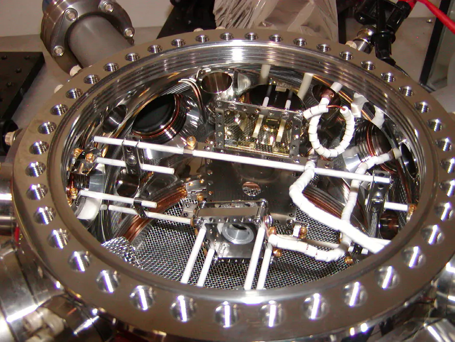 Looking down into a round stainless vacuum chamber through its open top flange: ceramic rods, beaded wires, a perforated floor and the cathode mount.
